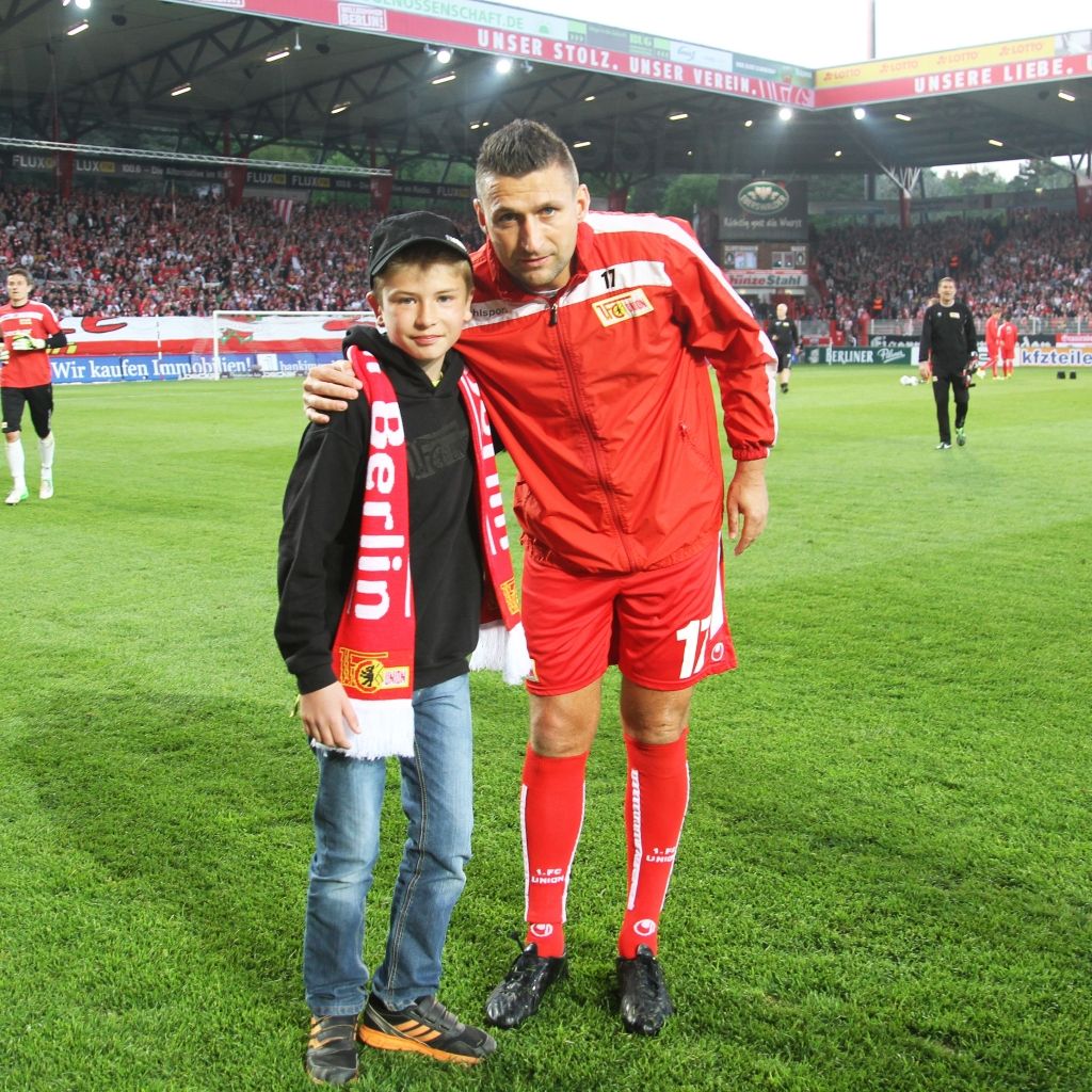 Ein Junge steht mit einem Fußballspieler auf dem Spielfeld, beide tragen Schals des Vereins. Im Hintergrund sieht man jubelnde Fans.