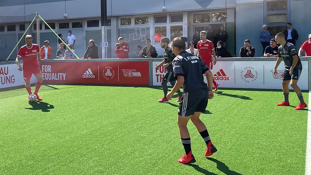 Fußballspieler trainieren auf einem Kunstrasenplatz, Zuschauer beobachten das Training im Hintergrund.