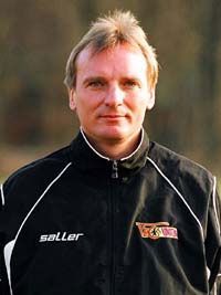 Ein Mann mit schulterlangen blond-grauen Haaren trägt eine Sportjacke und schaut direkt in die Kamera.