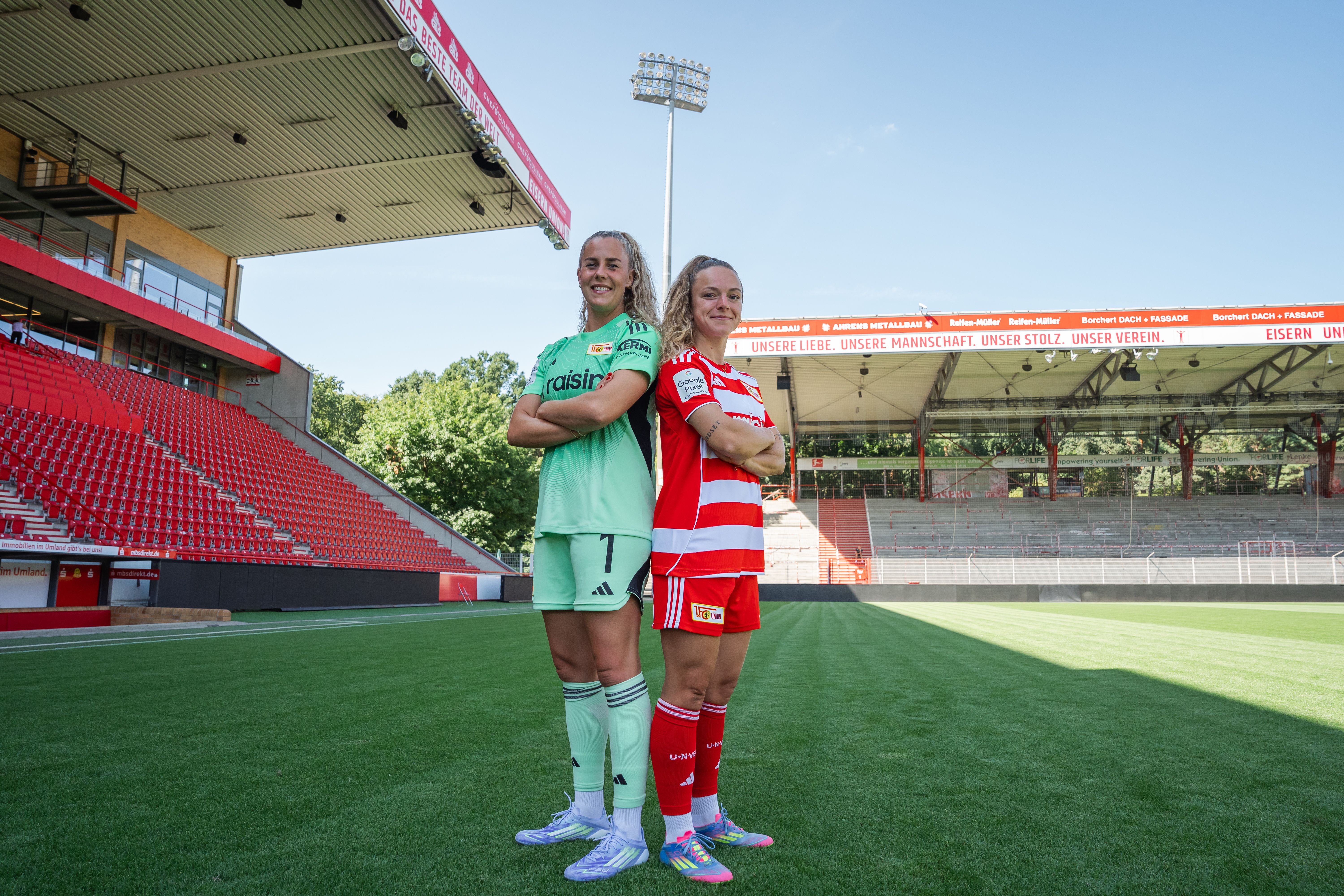 Two women in soccer jerseys stand on a grass field, seen from behind, with a stadium in the background.