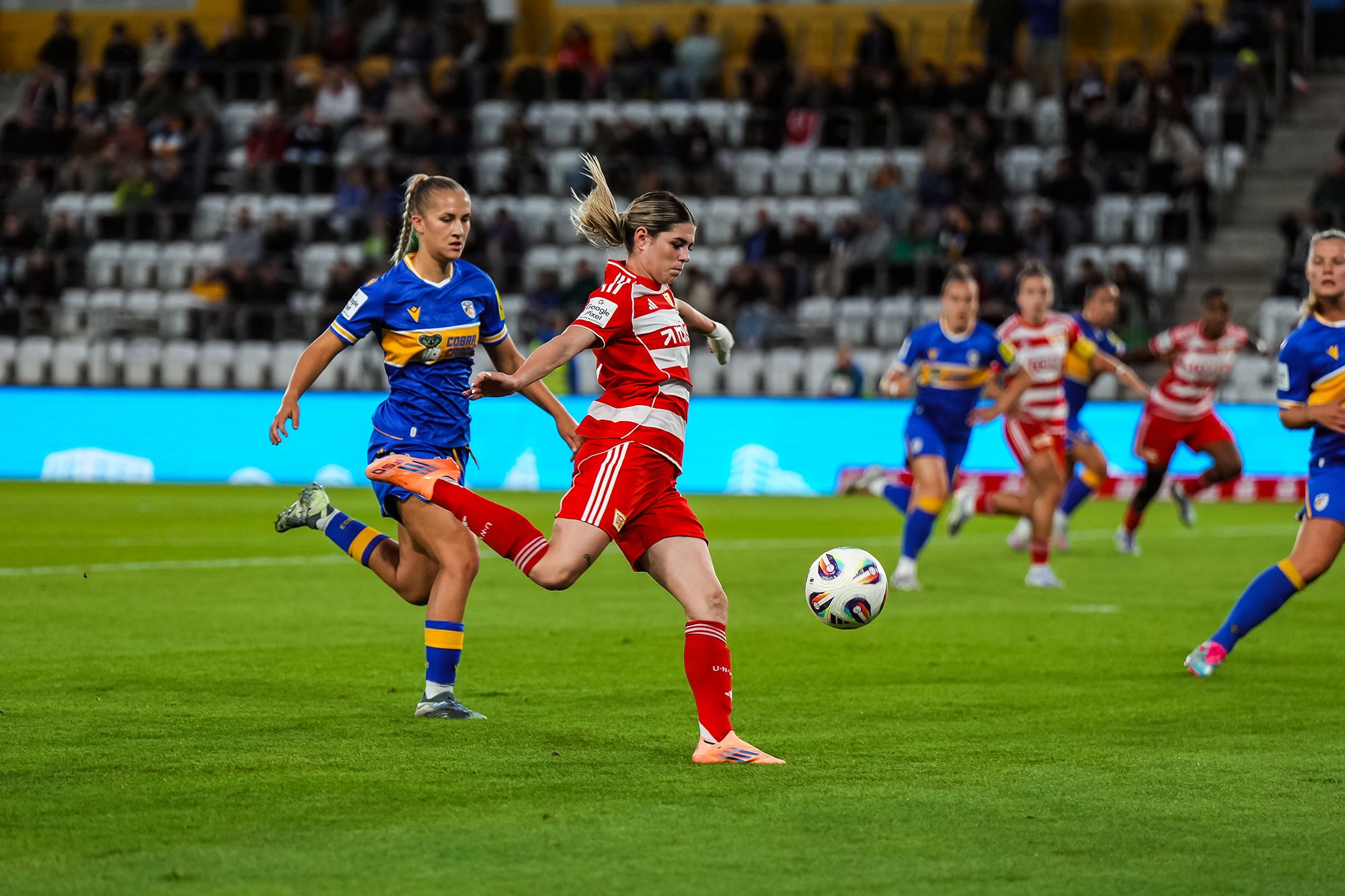 Spielszene eines Frauenfußballspiels, wo eine Spielerin in rotem Trikot den Ball schlägt, während eine Gegnerin in gelb-blauem Trikot nähert.