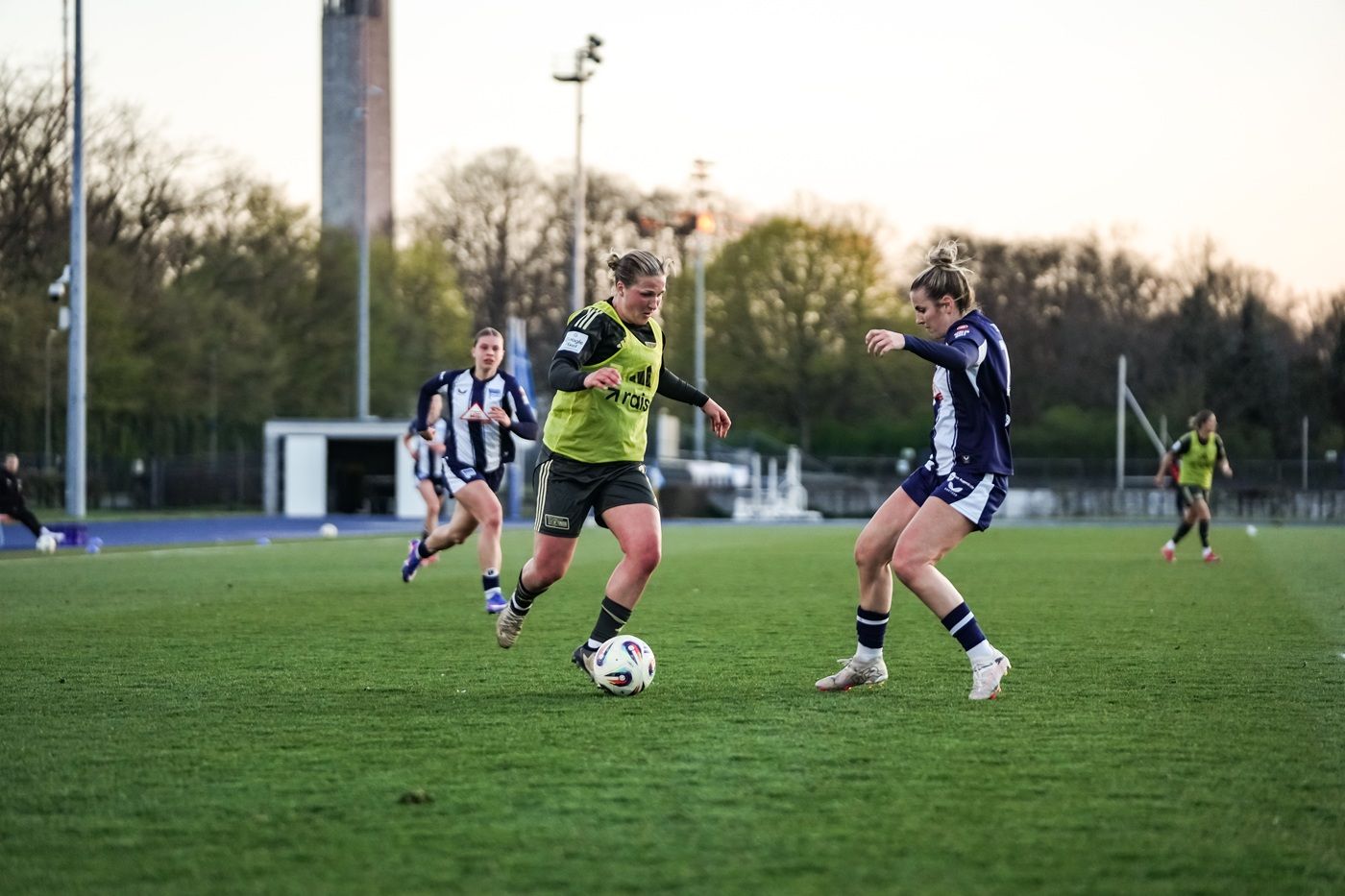Zwei Frauen im Fußballtrikot dribbeln mit einem Ball, während eine dritte Spielerin im Hintergrund läuft. Der Platz ist grün und beleuchtet.