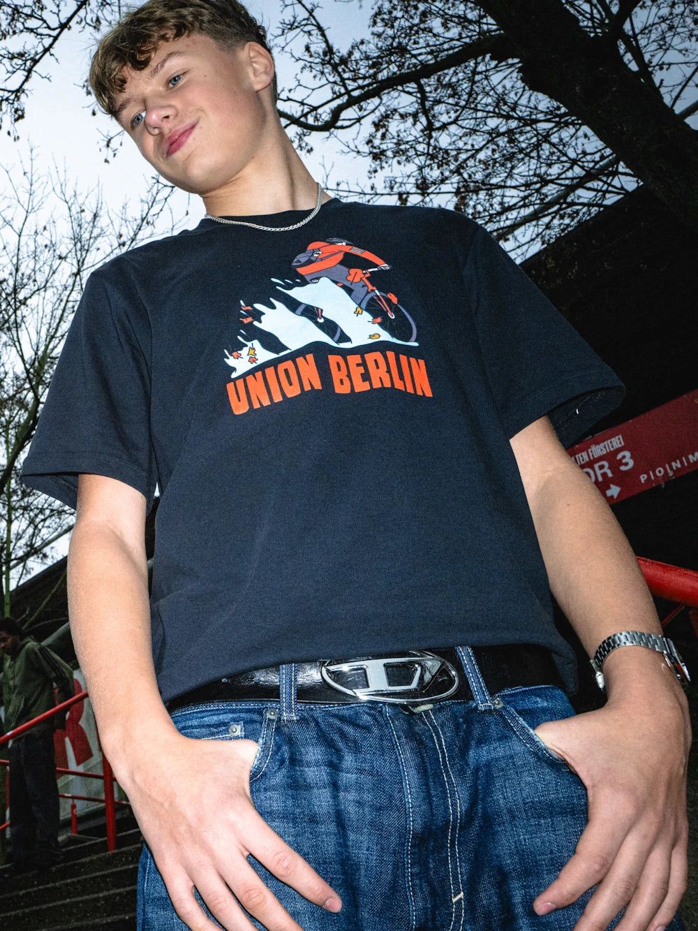 Junger Mann trägt ein T-Shirt mit dem Schriftzug "Union Berlin" und steht vor einer Szenerie mit Bäumen.