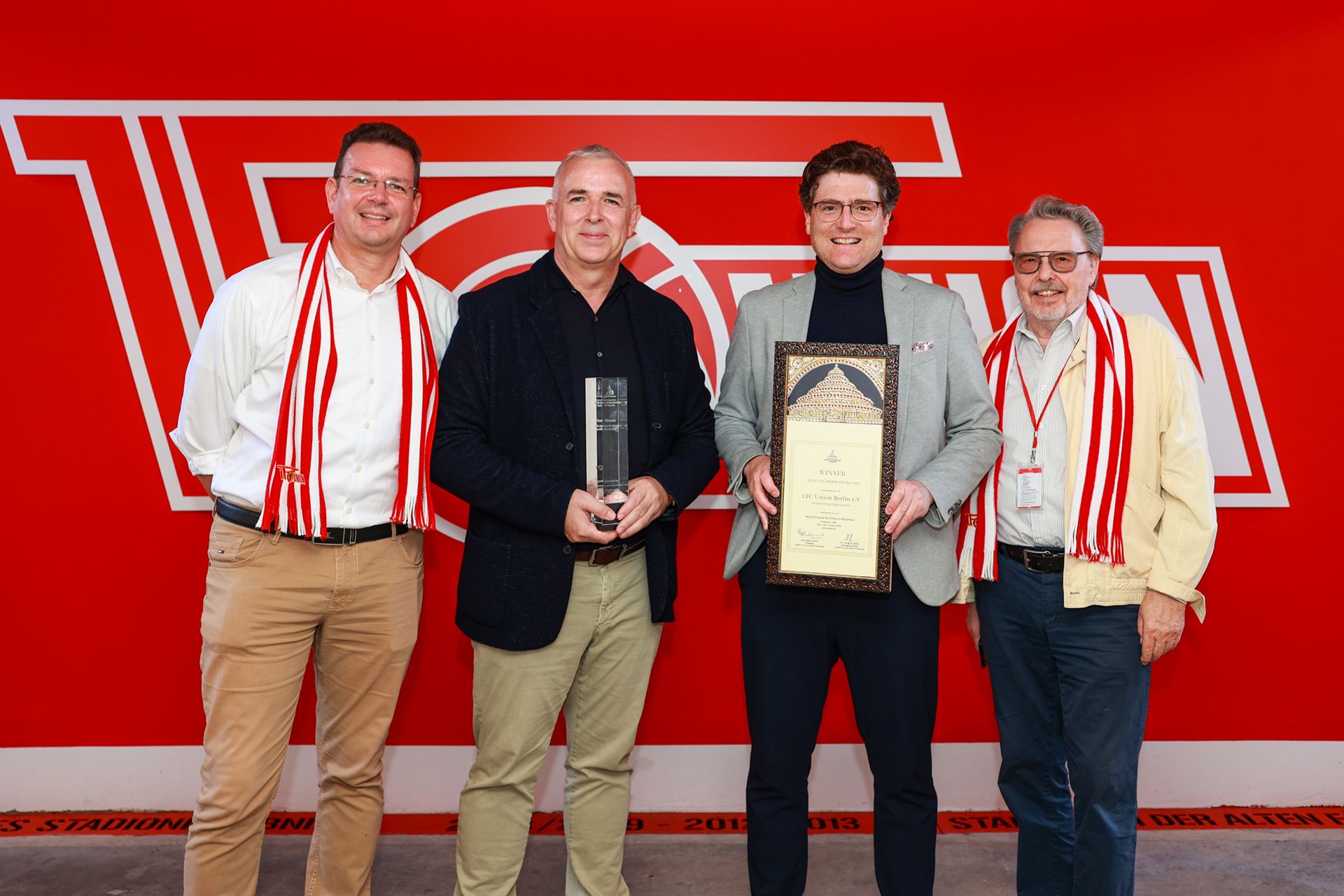 Vier Personen stehen zusammen vor einer roten Wand mit dem Logo eines Vereins, wobei zwei von ihnen eine Auszeichnung halten. Alle tragen einen Schal.