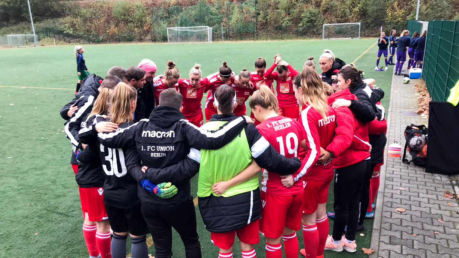 Eine Gruppe von Fußballerinnen trägt rote Trikots. Sie stehen zusammen in einem Kreis auf dem Platz.