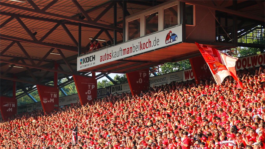 Eine volle Sporttribüne mit jubelnden Fans in roten T-Shirts und Fahnen, die ein Team unterstützen.