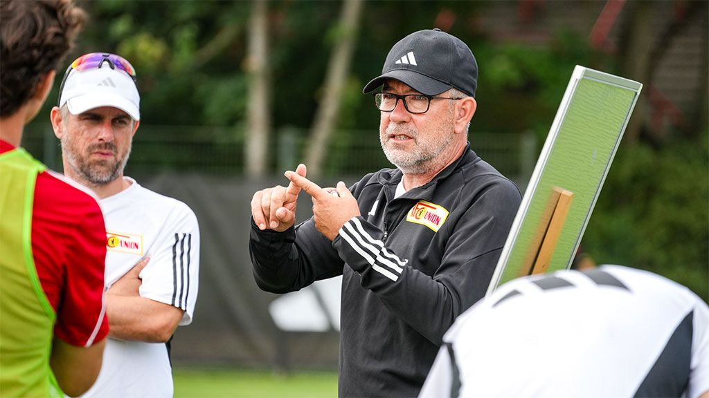 A coach explains tactics during a soccer drill, surrounded by players on a training field.