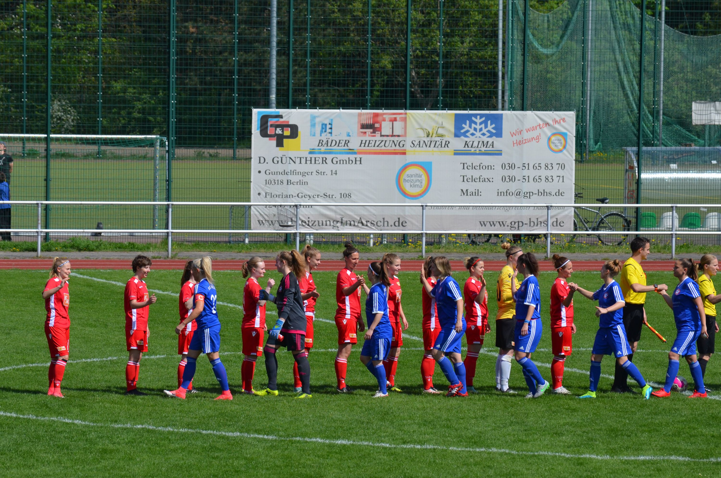 Zwei Fußballteams stehen auf einem Platz. Die Spieler tragen rote und blaue Trikots.
