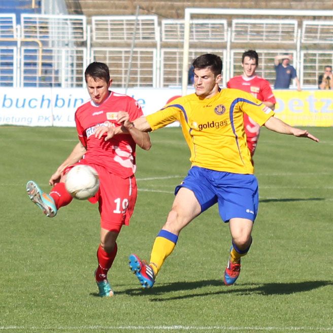 Zwei Fußballspieler in Aktion, einer mit rotem und einer mit gelbem Trikot, kämpfen um den Ball auf dem Feld.