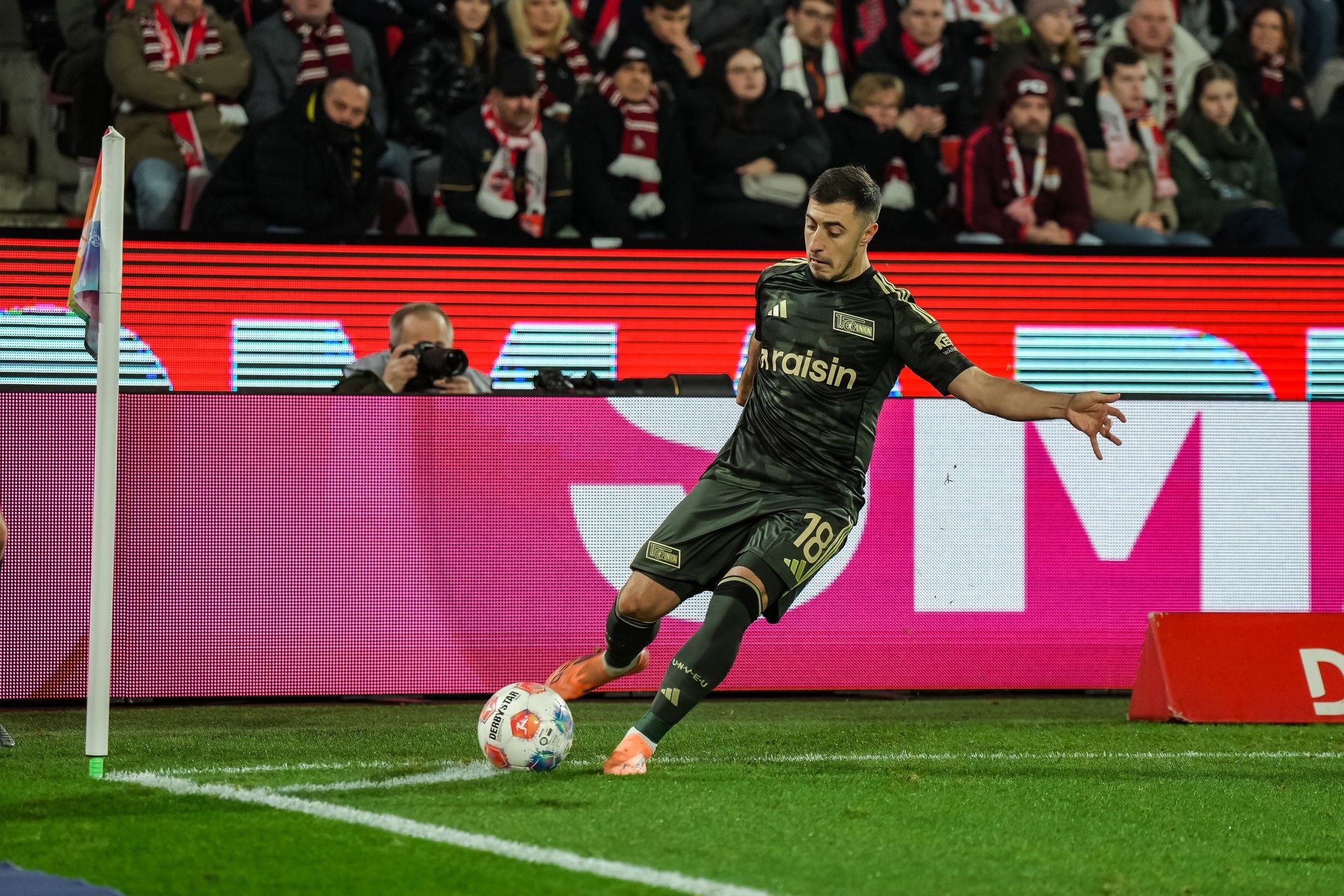 Spieler führt einen Eckball aus, während Zuschauer im Hintergrund auf das Spielgeschehen achten.