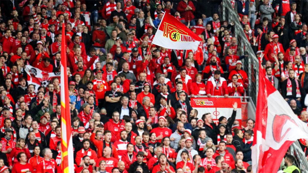 An enthusiastic crowd in red jerseys waving flags and cheering at a soccer game.