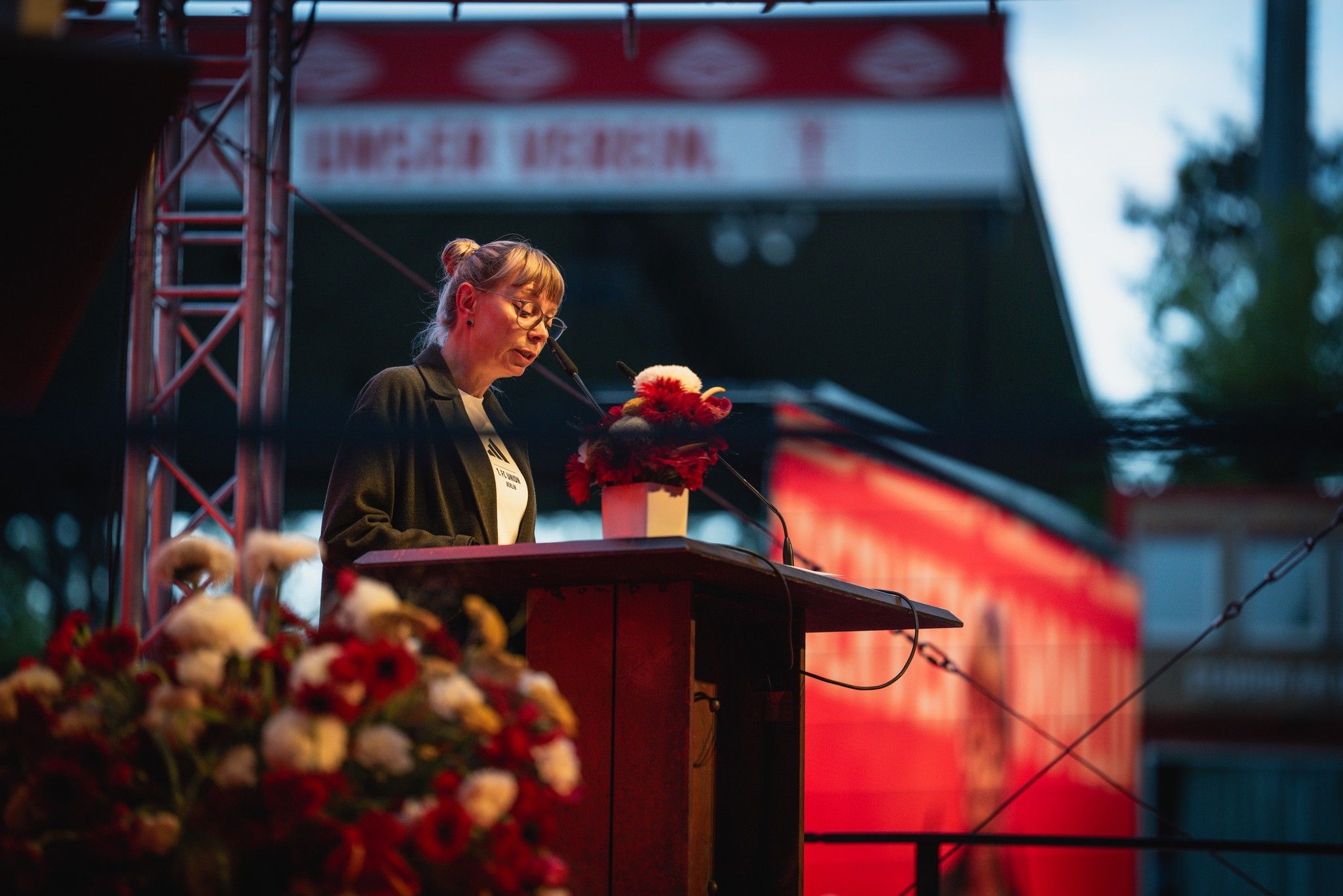 Frau spricht am Rednerpult vor einer großen, blumenarrangierten Kulisse bei einem Event. Rot und Weiß dominieren die Farben.
