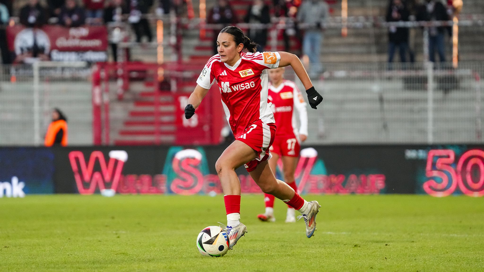 Spielerin in rotem Trikot dribbelt wartend mit dem Ball auf dem Fußballfeld. Zuschauer im Hintergrund.