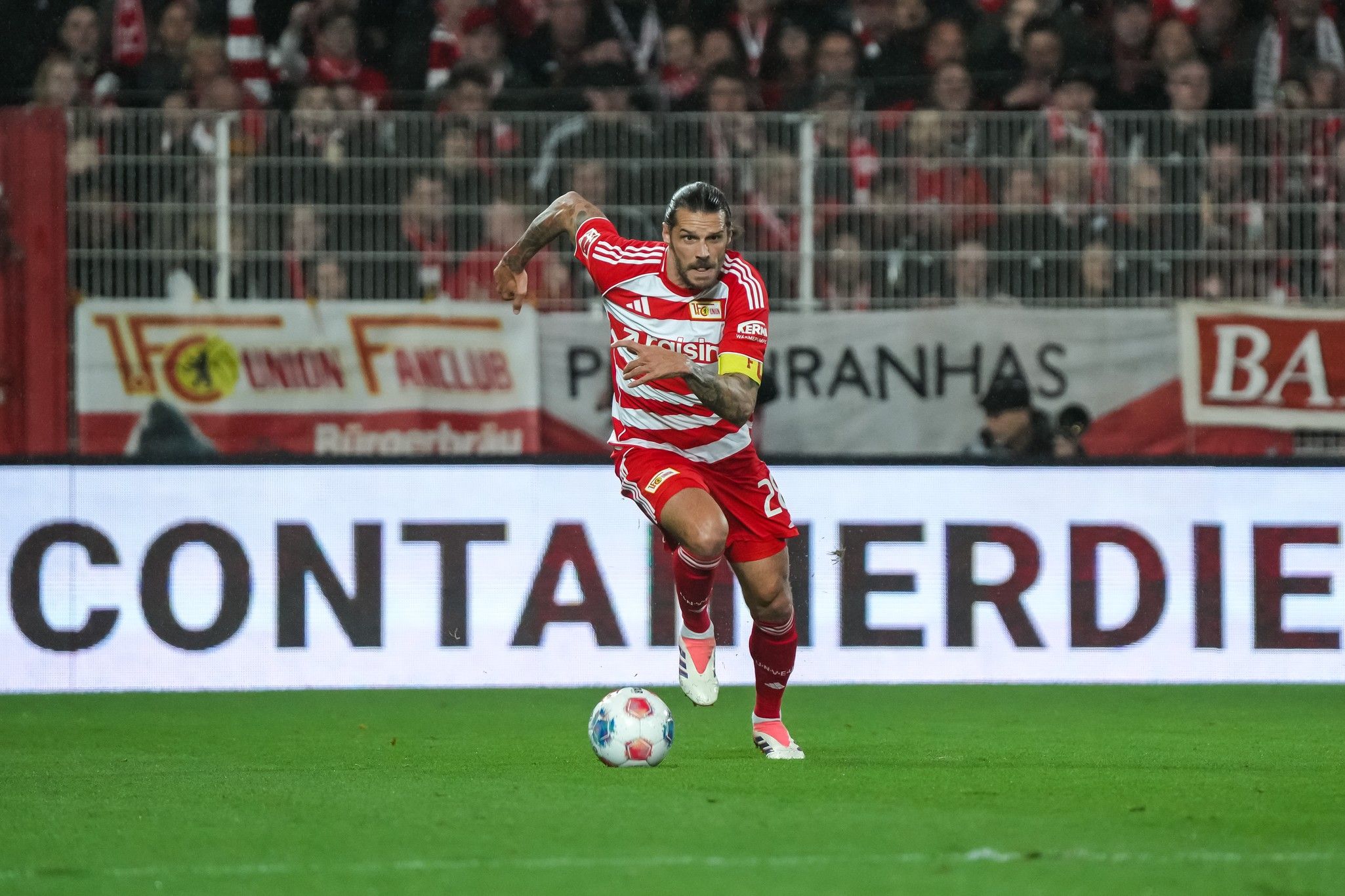 Ein Fußballspieler mit Kapitänsbinde läuft während eines Spiels, im Hintergrund sind jubelnde Fans und Werbung zu sehen.