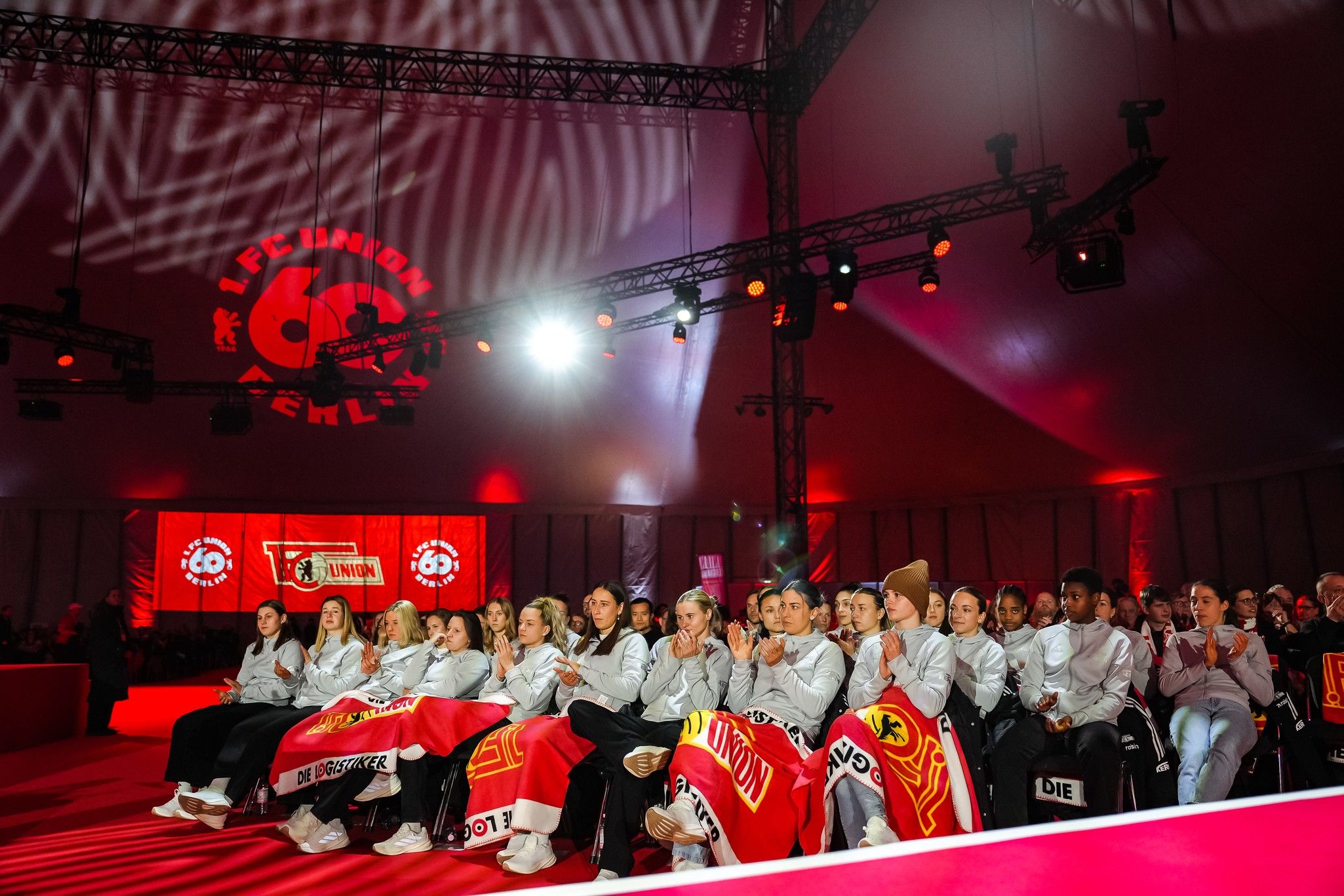 Eine Gruppe von Personen sitzt in einem festlichen Zelt, umgeben von rotem Licht und Dekorationen mit dem Logo von Union Berlin.