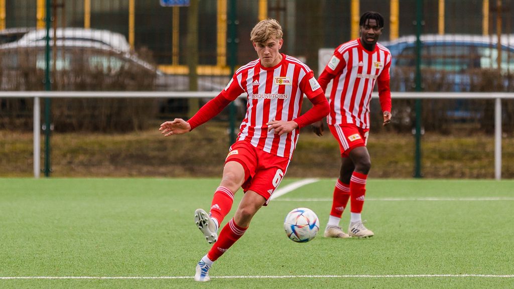 Ein Fußballspieler in rot-weiß gestreiftem Trikot tritt den Ball, während ein Mitspieler im Hintergrund aufmerksam zusieht.