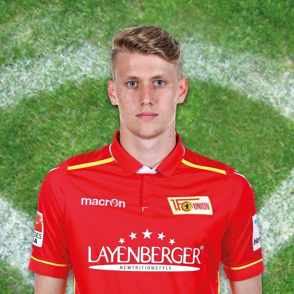 Portrait eines jungen Fußballspielers in rotem Trikot mit dem Logo des 1. FC Union Berlin vor einem grünen Hintergrund.