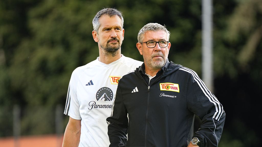 Zwei Männer in Sportbekleidung stehen auf einem Trainingsplatz und beobachten das Geschehen.