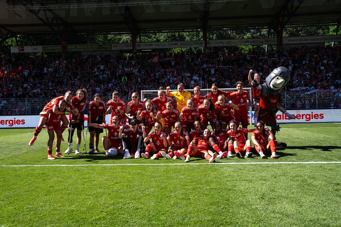 Eine Fußballmannschaft in roten Trikots posiert jubelnd auf dem Rasen, umgeben von Fans im Stadion.