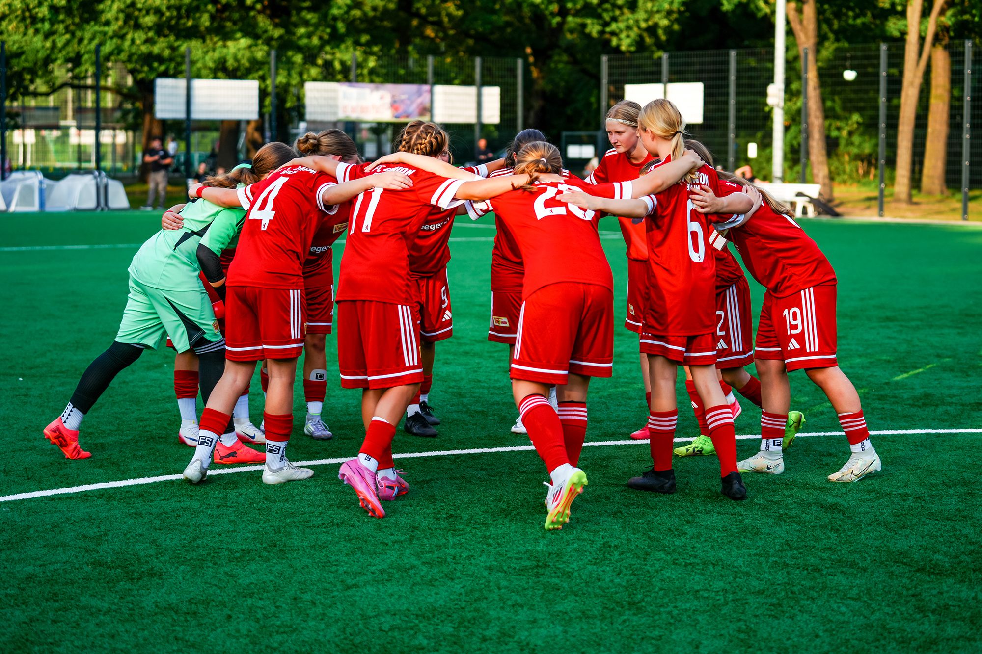 Eine Gruppe von Mädchen in roten Fußballtrikots umarmt sich auf einem Sportplatz.