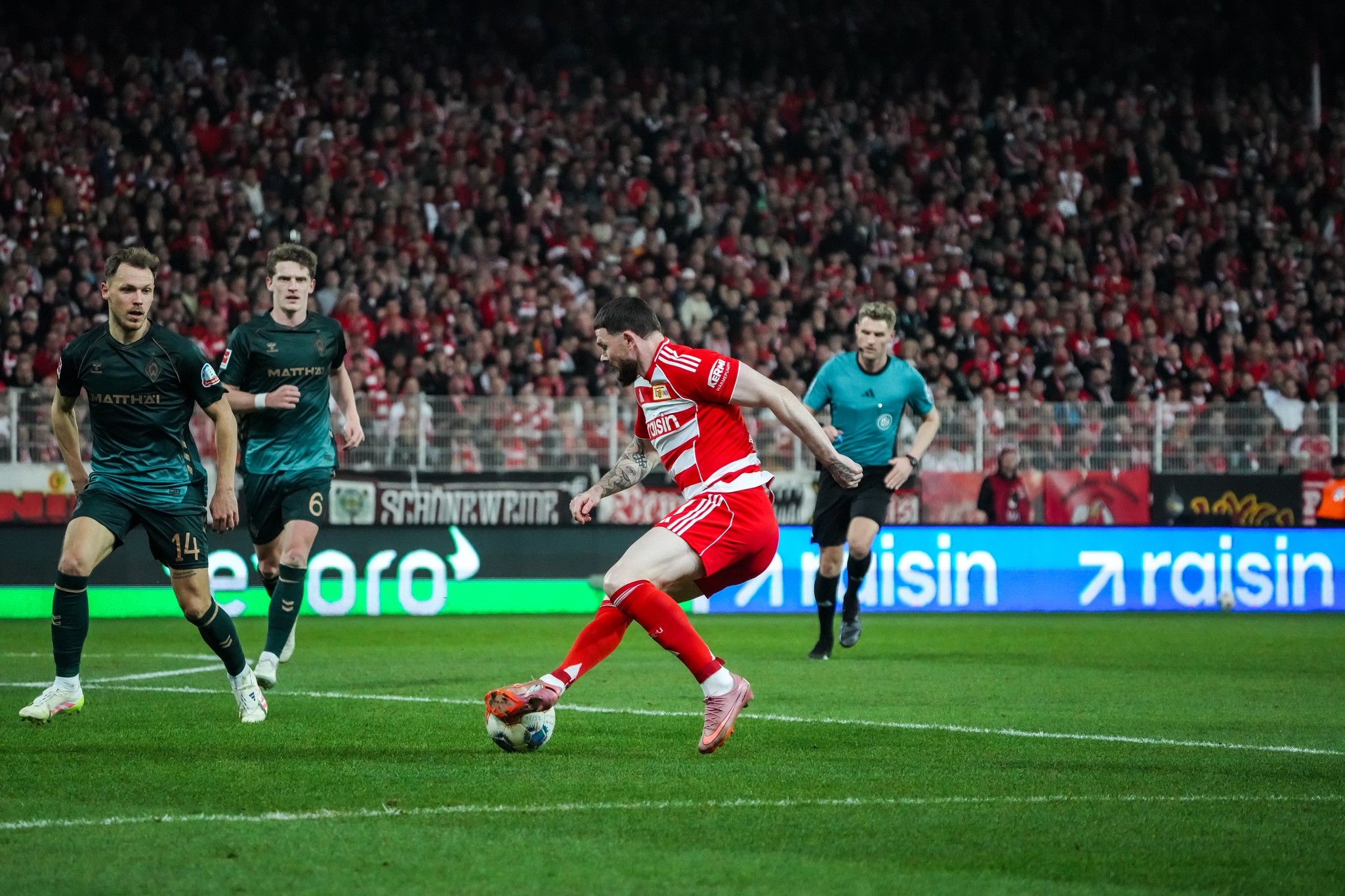 Ein Fußballspieler in rotem Trikot dribbelt den Ball, während seine Gegner in dunklen Trikots ihn beobachten. Fans im Hintergrund.