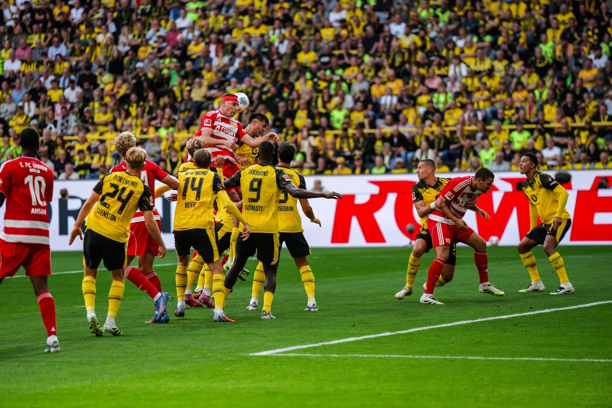 Spielszene im Fußballstadion: Spieler springen hoch, um den Ball während einer Ecke zu erreichen, umgeben von einer belebten Zuschauertribüne.
