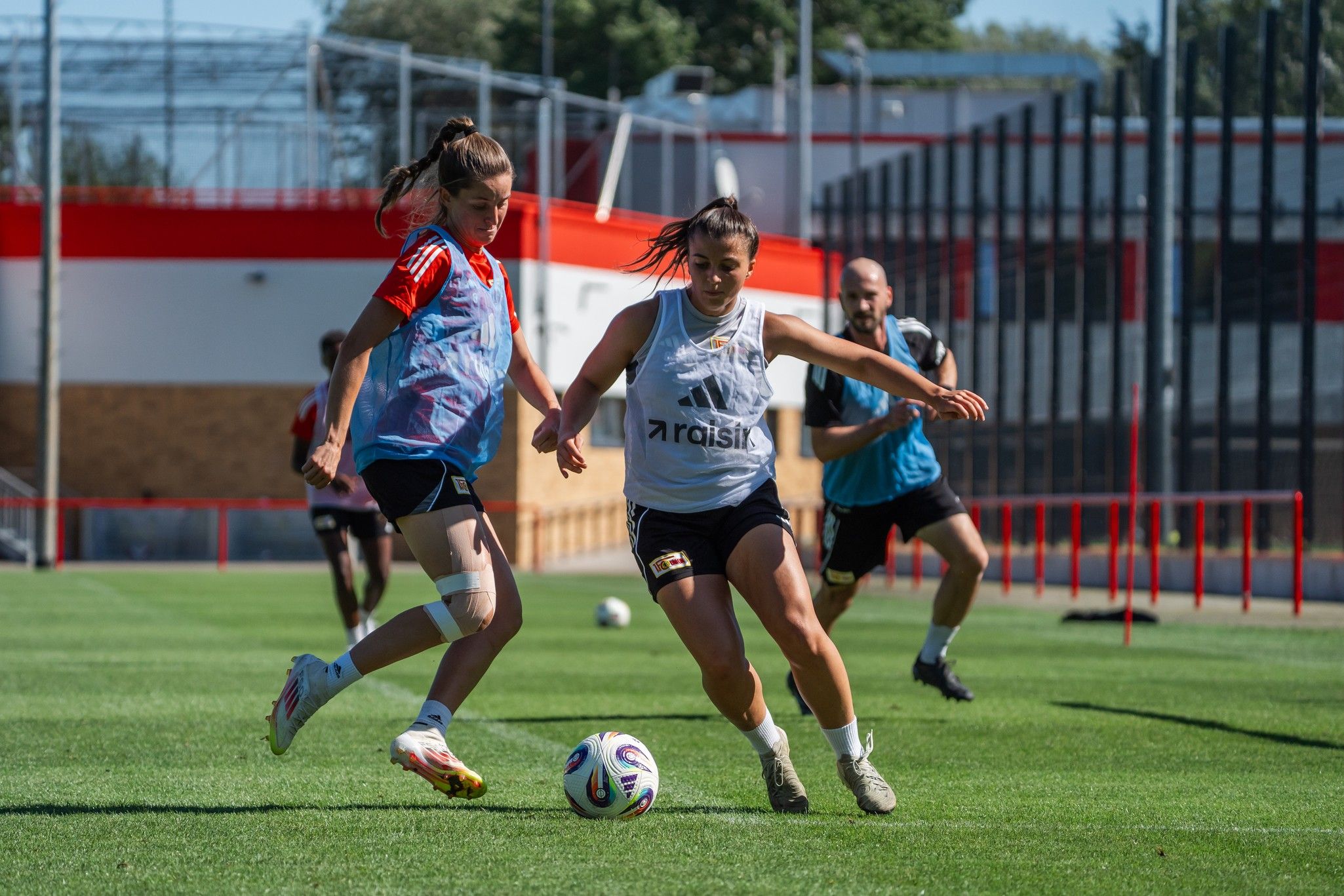 Zwei Frauen spielen auf einem Fußballfeld, während ein Mann im Hintergrund zuschaut. Sonnenschein und grünes Gras sind sichtbar.