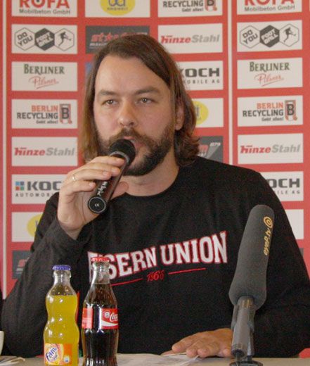 Mann mit Bart spricht am Mikrofon, trägt ein schwarz-rot gestreiftes Sweatshirt, neben ihm stehen Getränke auf einem Tisch.