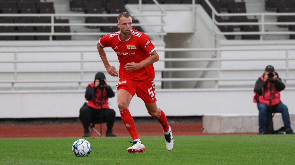 A soccer player in a red jersey runs across the field while photographers stand in the background.