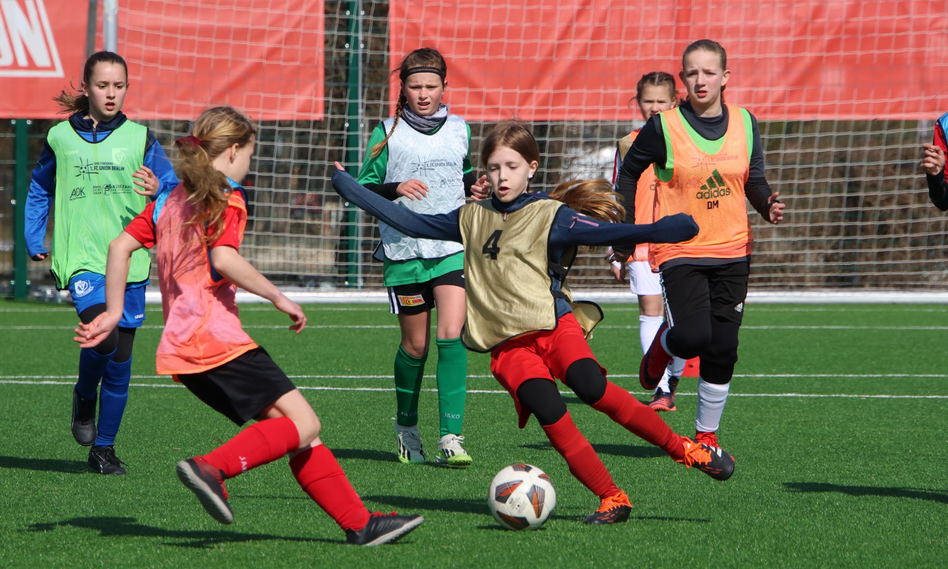 Mädchen spielt Fußball auf einem Rasenfeld, drei Spielerinnen in verschiedenen Trikots sind aktiv.