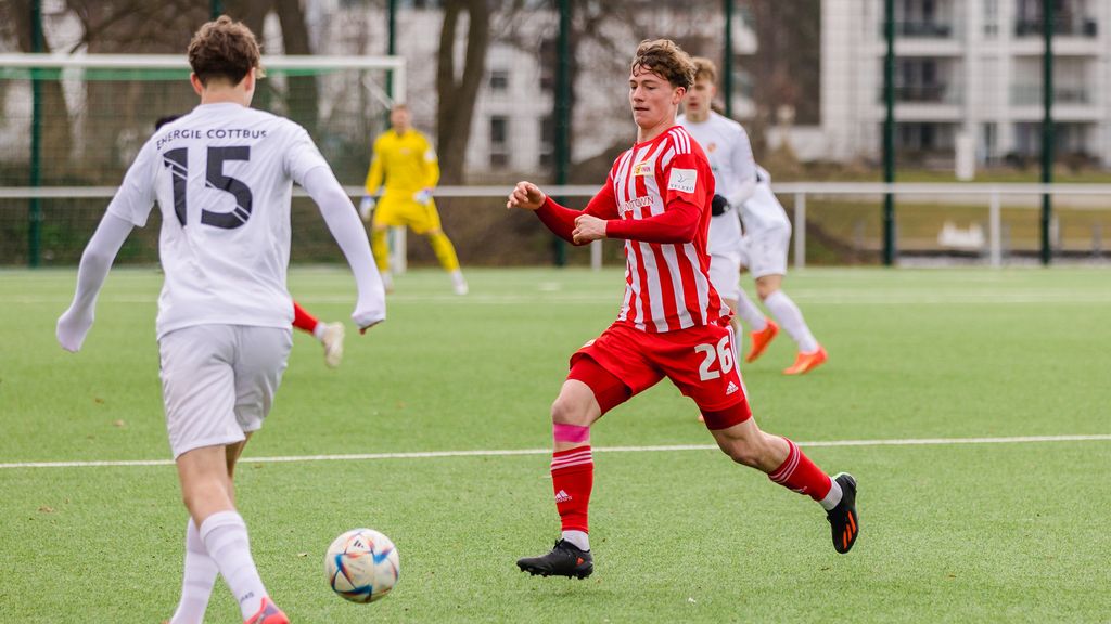 Ein Fußballspieler in rotem Trikot mit der Nummer 26 dribbelt den Ball auf einem Kunstrasenplatz, während ein Spieler in Weiß ihm entgegenläuft.
