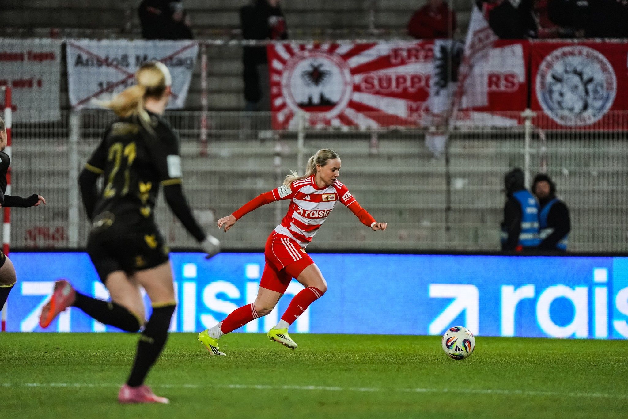 Eine Fußballspielerin in rot-weißer Kleidung sprintet mit dem Ball, während eine Torhüterin im Hintergrund steht. Fanbanner sind sichtbar.