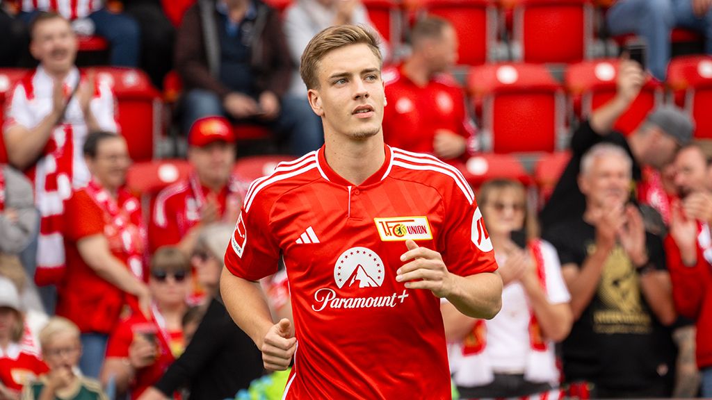 Ein Fußballspieler in roter Uniform läuft fröhlich auf das Spielfeld, während Fans in der Tribüne applaudieren.