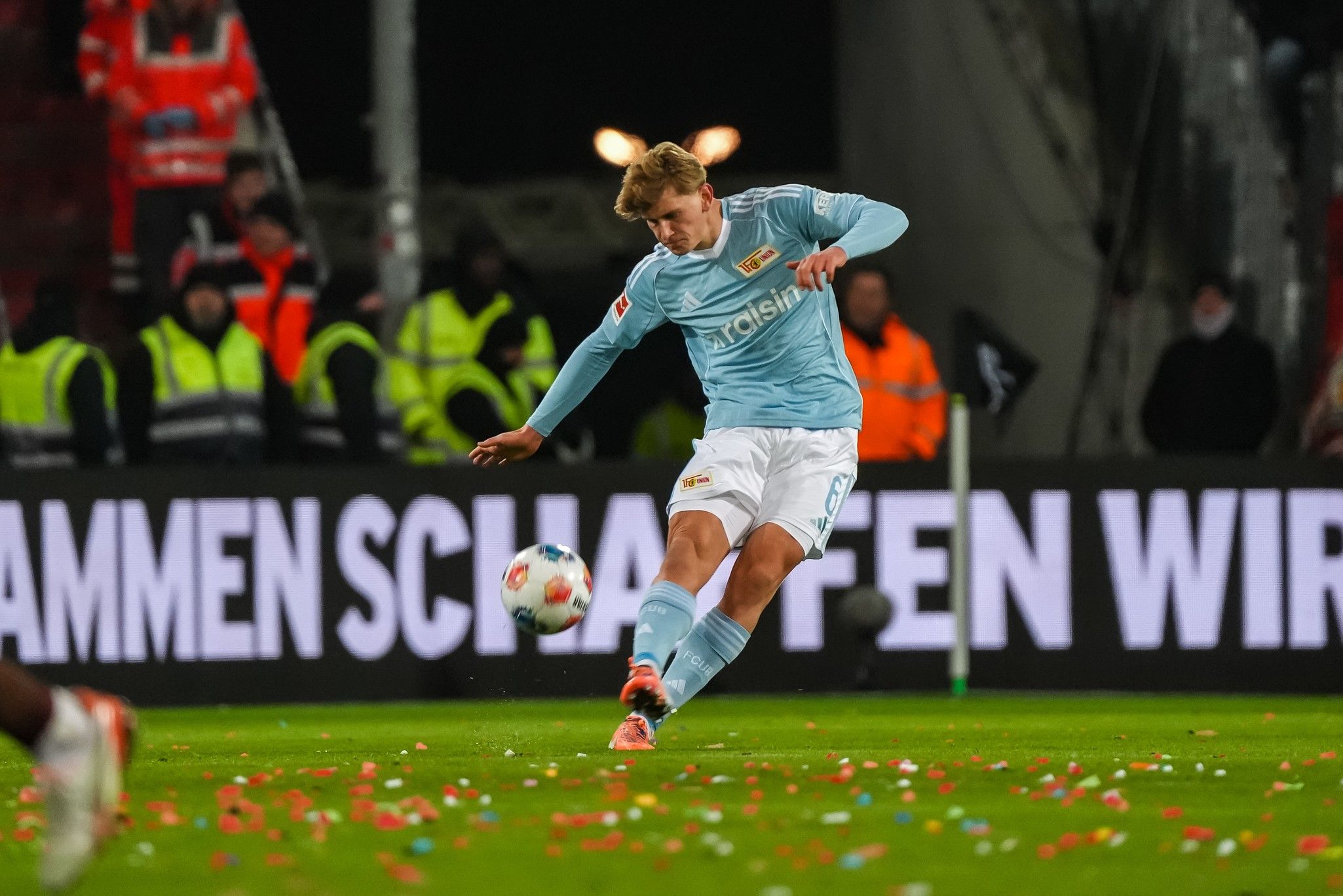 Ein Spieler in blauer Mannschaftskleidung schießt den Ball auf einem Fußballfeld, umgeben von jubelnden Fans und Konfetti.