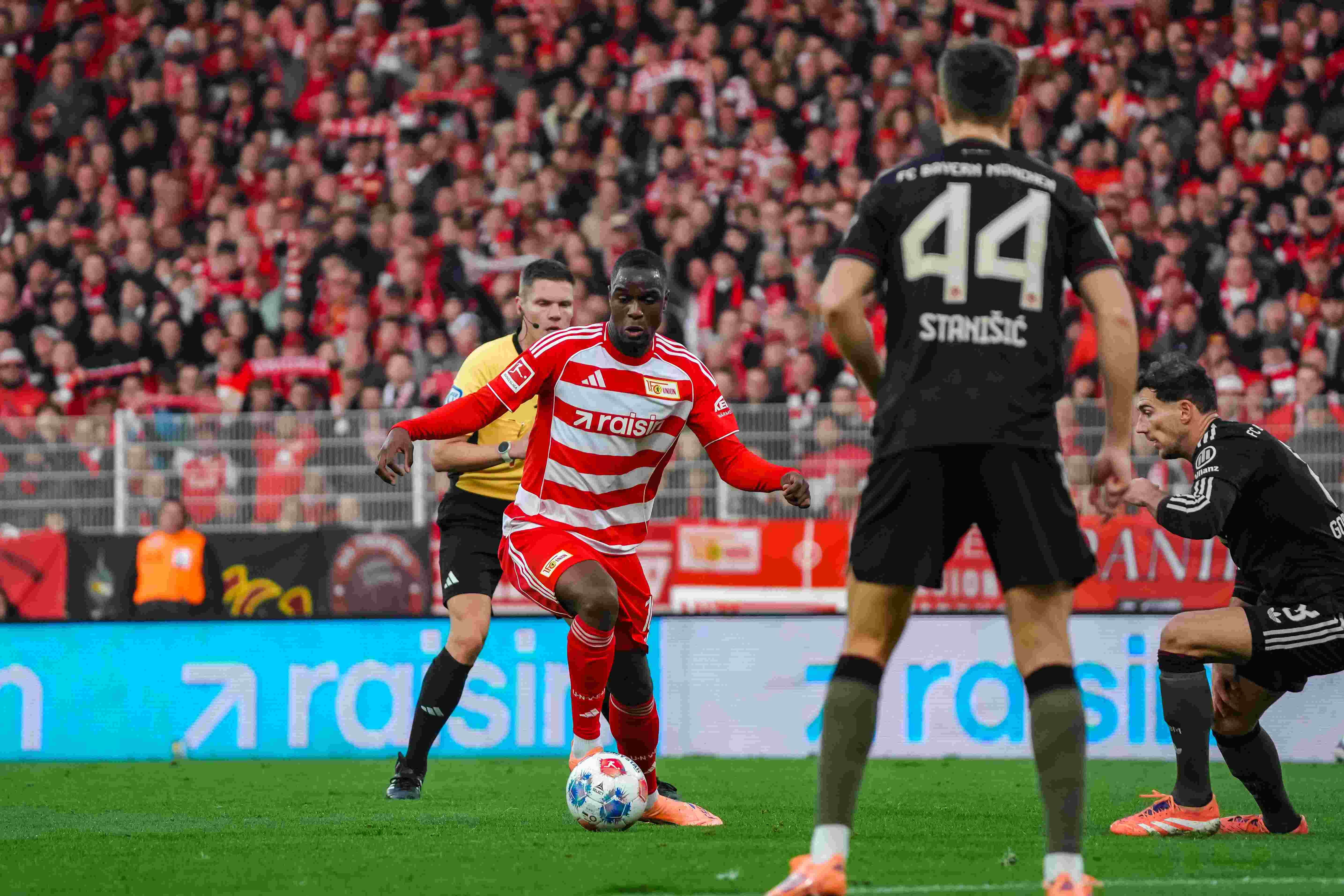 Ein Fußballspieler trägt ein rotes Trikot. Er tritt den Ball. Im Hintergrund sind zwei Gegner und ein Schiedsrichter.