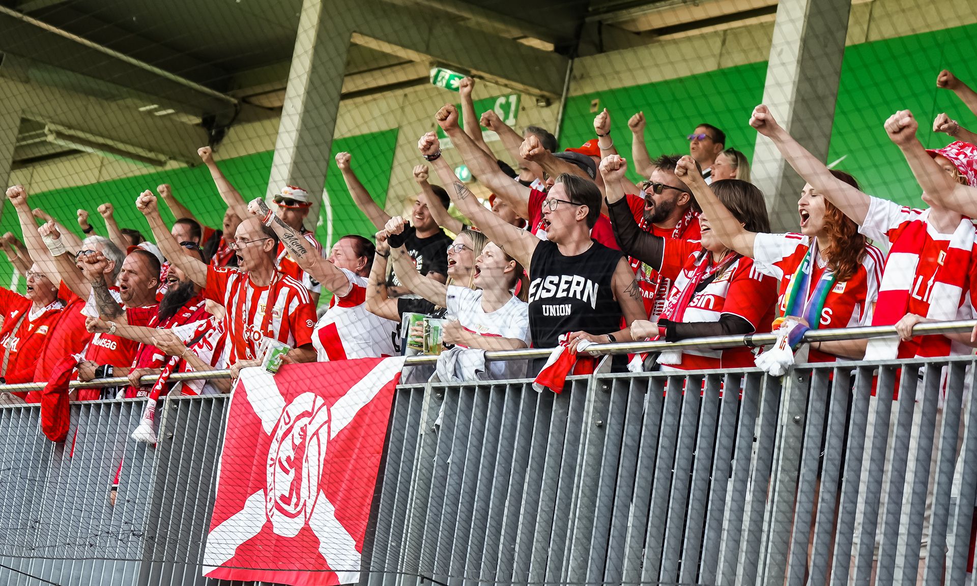 Eine Gruppe von jubelnden Fans in rot-weißen Trikots, die mit erhobenen Fäusten und Fahnen vor einem Stadion stehen.