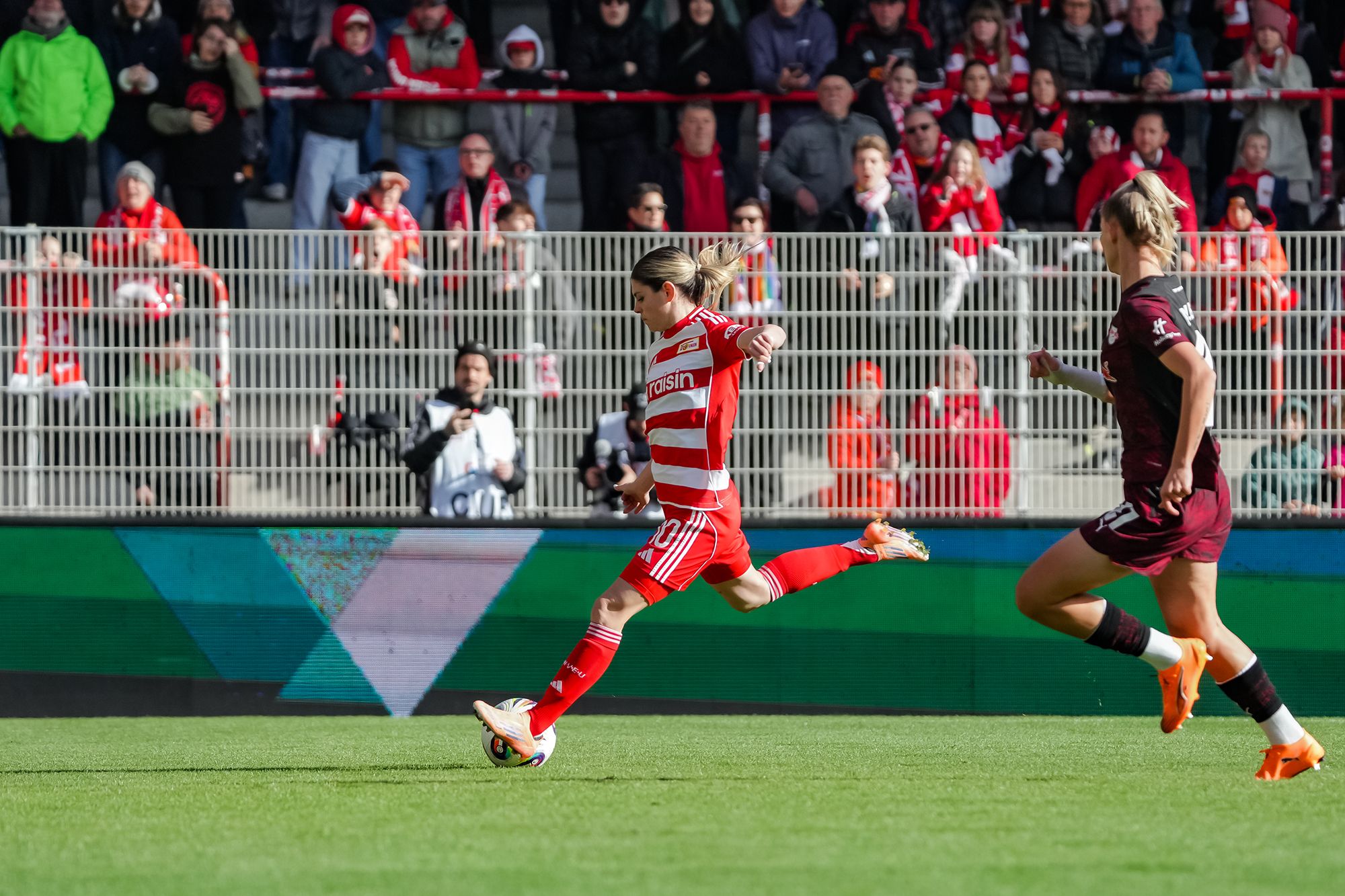 Eine Spielerin in rot-weißer Sportkleidung sprintet mit dem Ball, während ein Gegenspieler in dunkler Kleidung sie verfolgt.
