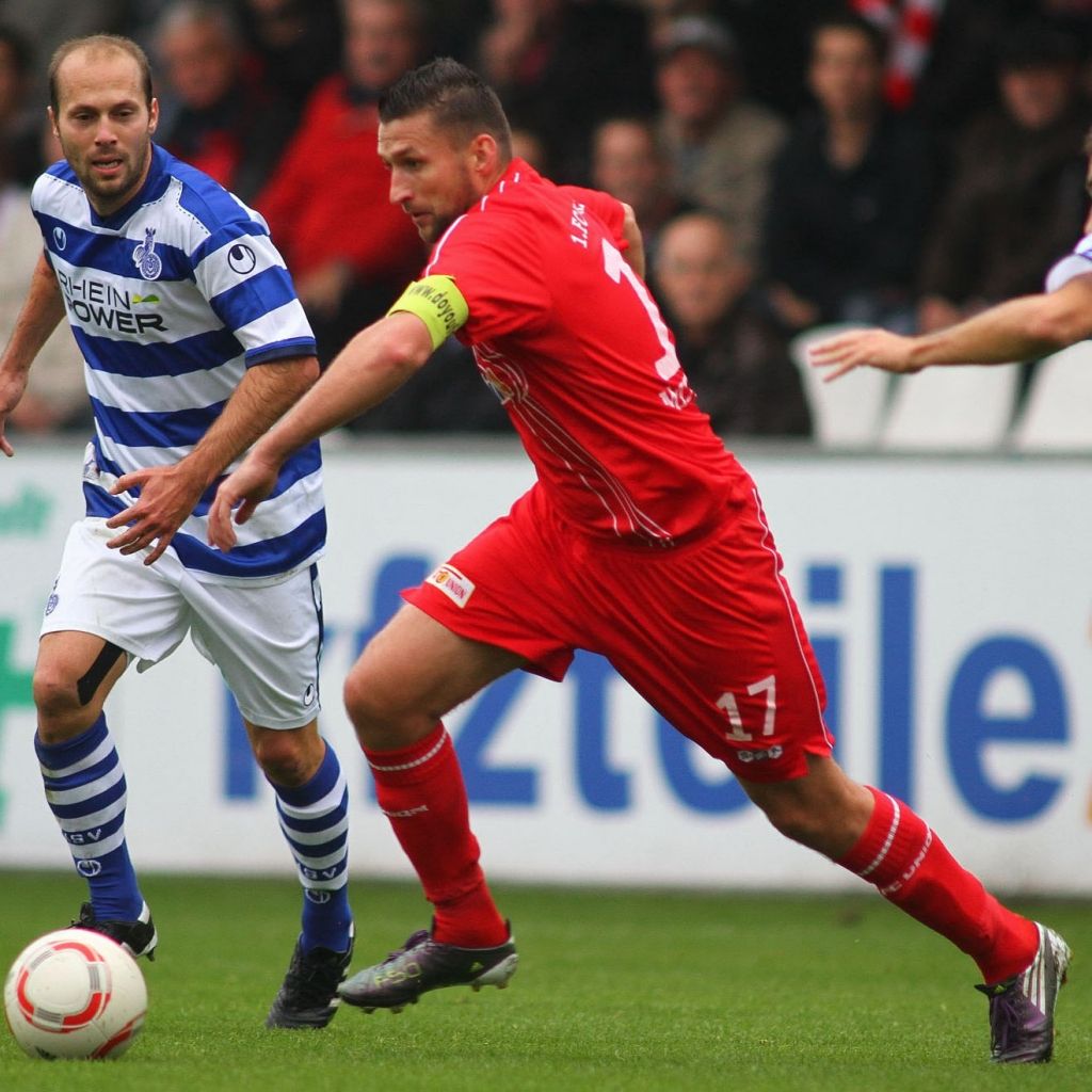 Zwei Fußballspieler auf dem Feld: einer in gestreiftem Trikot mit Ball, der andere in rotem Trikot, auf dem Weg zur Balleroberung.