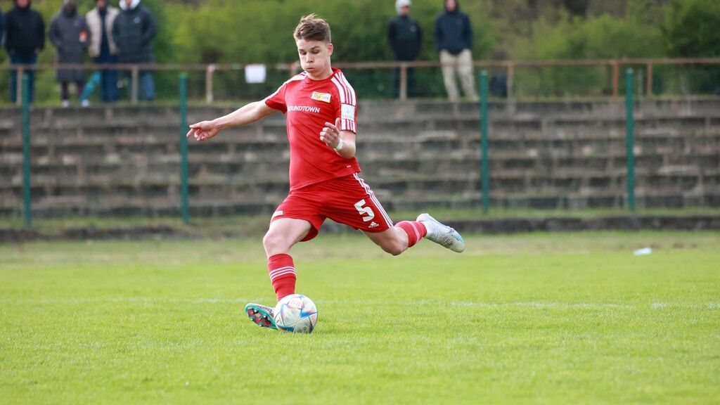 Ein junger Fußballspieler in rotem Trikot führt einen Schuss auf dem Spielfeld aus, während Zuschauer im Hintergrund stehen.