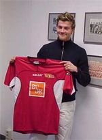 Ein lächelnder Mann hält ein rotes Fußballtrikot mit einem Logo in der Mitte in einem Raum mit Fotos an der Wand.