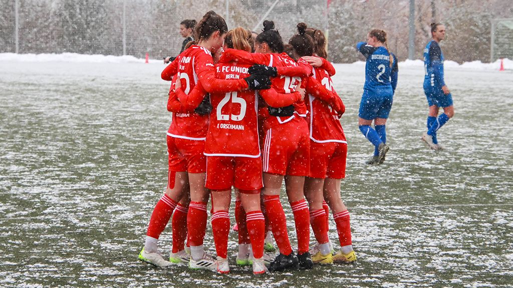 Eine Gruppe von Fußballspielerinnen in roten Trikots umarmt sich. Sie stehen auf einem Spielfeld mit Schnee. Im Hintergrund sind Spielerinnen in blauen Trikots.