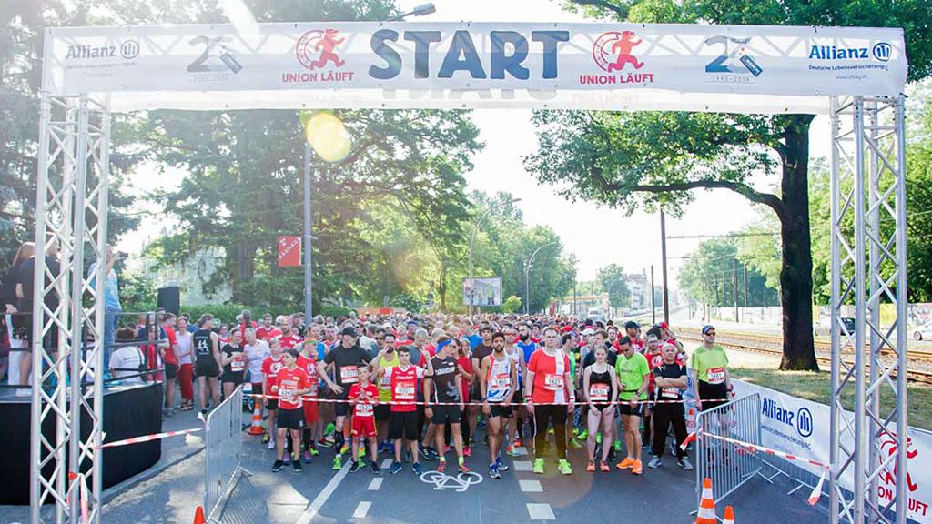 Startlinie eines Laufwettbewerbs mit einer großen Gruppe von Teilnehmern, die in rot und schwarz gekleidet sind, vor einem Banner.