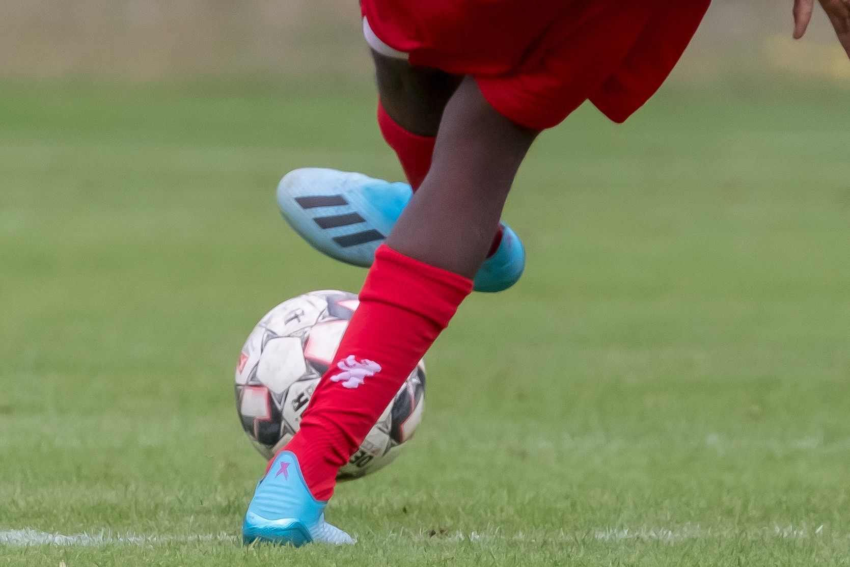 Ein Fußballspieler mit roten Socken und blauen Schuhen tritt einen Fußball auf einem grünen Rasenfeld.