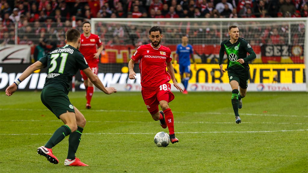 A player in a red jersey dribbles the ball during a soccer match, accompanied by two players in green jerseys.
