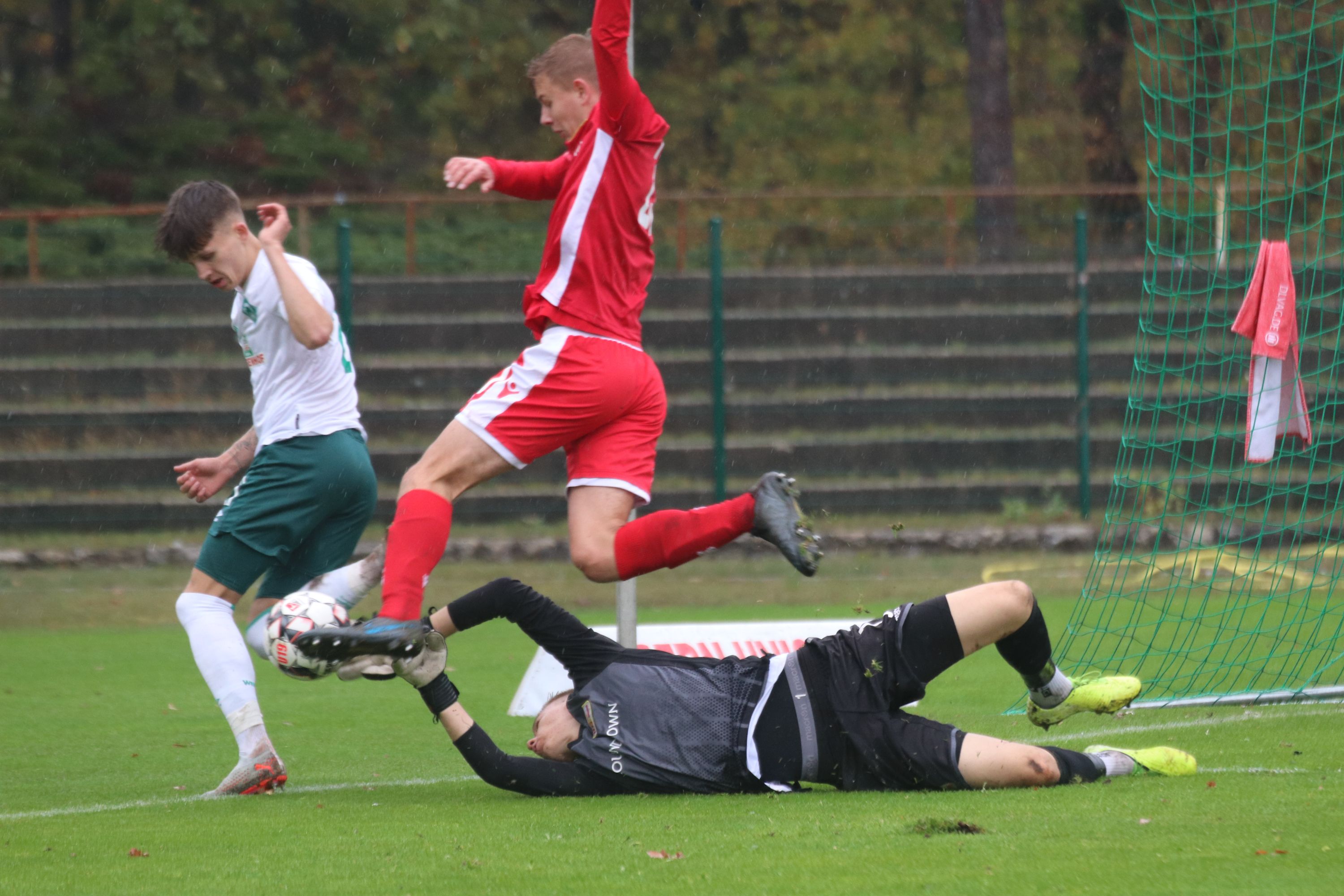 Fußballspiel mit einem Torwart, der den Ball hält, während ein Spieler in roter Kleidung ihn herausfordert.