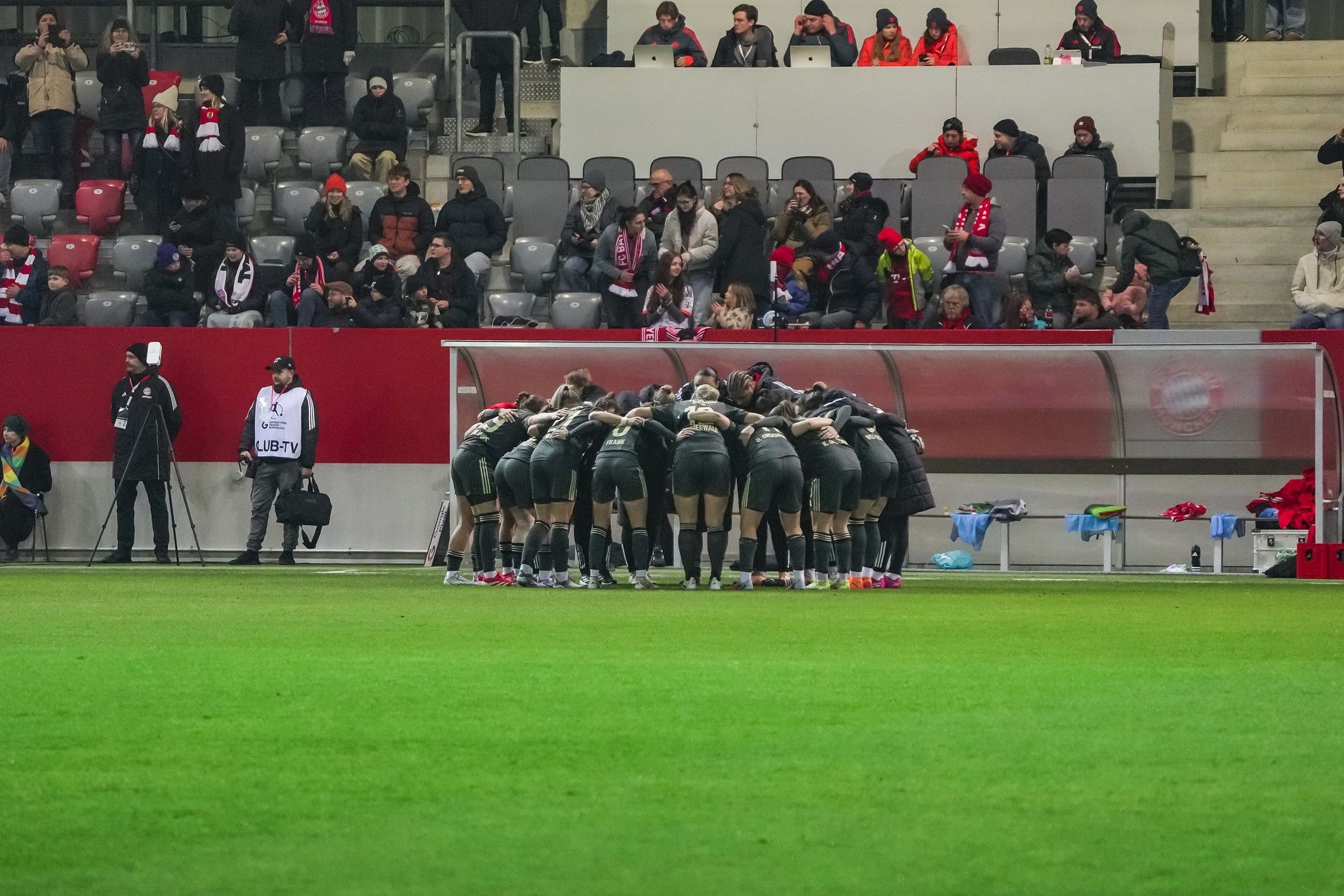 Eine Gruppe von Sportlern in schwarzen Trikots steht in einem Kreis auf dem Spielfeld, während Zuschauer im Hintergrund sitzen.