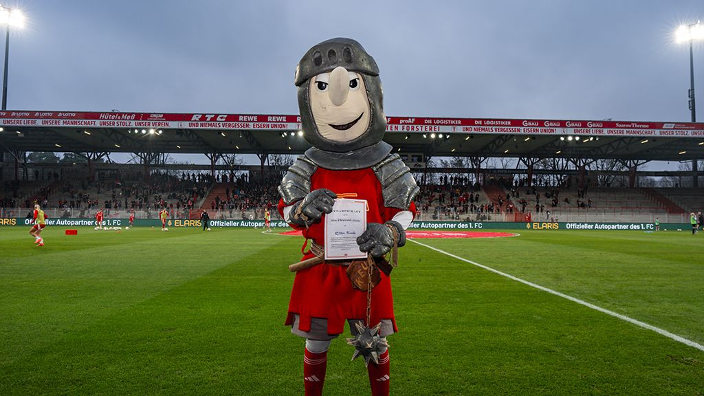 A mascot in knight costume holds a certificate, standing on a soccer field with empty stands in the background.