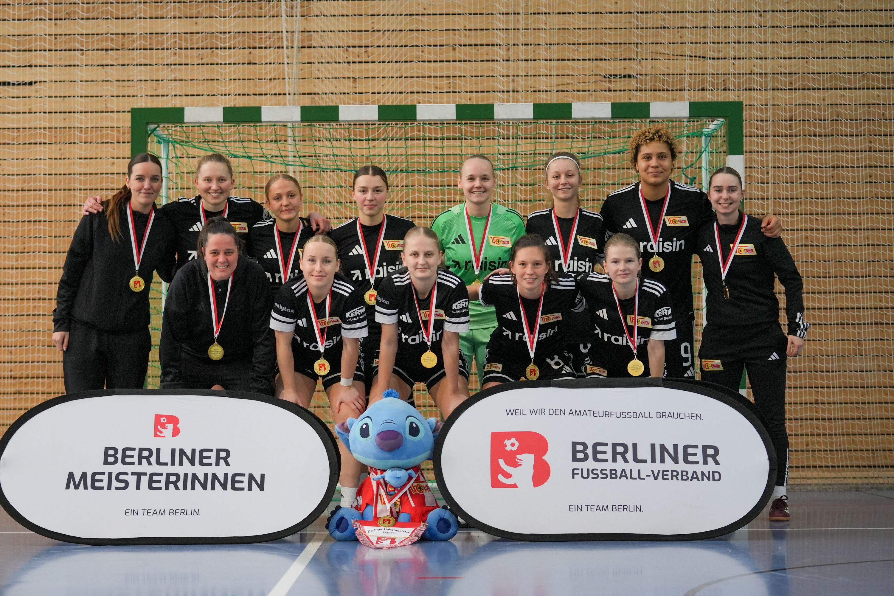 Die Siegerinnen einer Sportveranstaltung posieren in Trikots mit Medaillen vor einem Tor, umgeben von zwei Bannern.