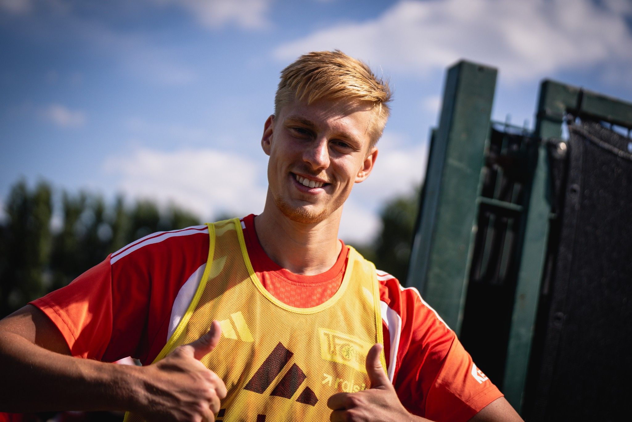 Ein junger Mann mit blonden Haaren in einem roten Trikot und gelber Trainingsweste zeigt Daumen nach oben.