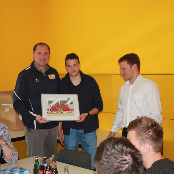 Drei Männer in einem Raum mit gelber Wand, einer hält ein gerahmtes Bild, während die anderen zuschauen. Getränke auf dem Tisch.